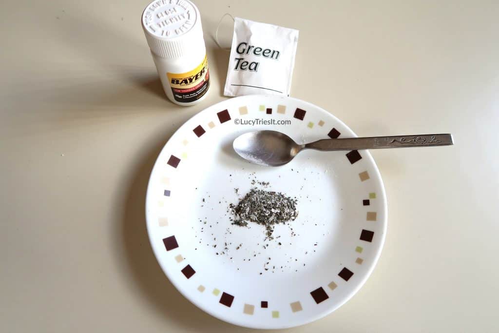 Crushed aspirin and green tea leaves mixed together on a plate with aspirin bottle, spoon, and green tea bag on the side.