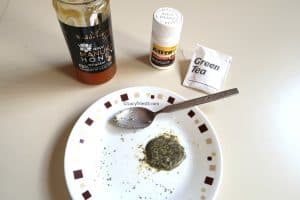 A homemade green tea and honey face mask mixture on a plate with honey jar, aspirin bottle, and green tea bag on the side and spoon sitting next to the mask.