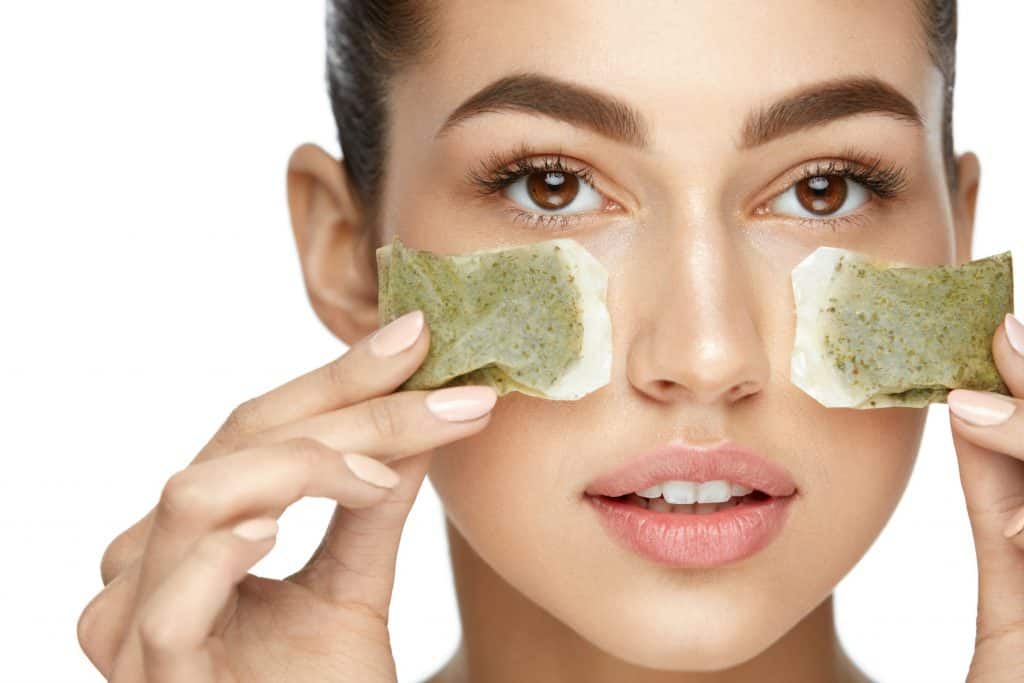 Young Woman With Natural Facial Makeup Holding Green Tea Bags On White Background for article answering the question, is green tea good for acne?